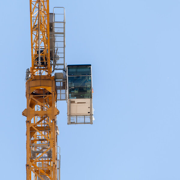 Fragment of yellow hoisting crane with cab on blue sky background.