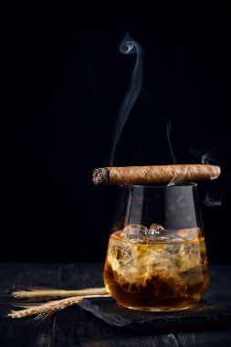 Bir bardak viski ya da üzerinde buz küpleri olan bir konyak ve üzerinde sigara içilen siyah ahşap bir arka plan.