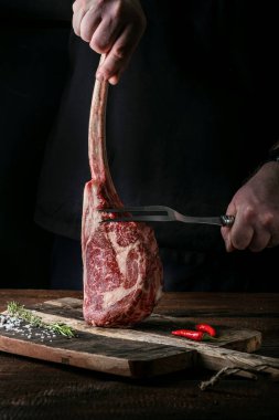 Şefin elleri çiğ bir Tomahawk biftek ve et çatalını doğrama tahtasının üzerinde tutuyor.