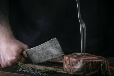 Bir kasap, siyah bir arka planda tahta bir kesme tahtasıyla kemiğe Tomahawk bifteği keser.