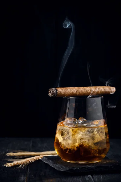 A glass of whiskey or cognac with ice cubes and a Smoking cigar lying on top, black wooden background