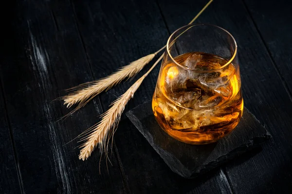 A glass of whiskey or Bourbon with ice cubes on a black wooden table with wheat grains