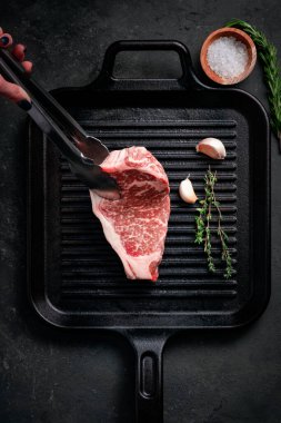 Çiğ Striploin bifteği ızgara tavasına maşa, sarımsak ve kekik konur.