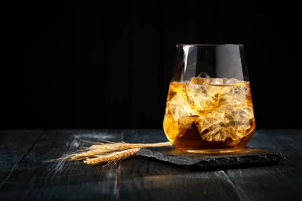 A glass of whiskey or Bourbon with ice cubes on a black wooden table with wheat grains