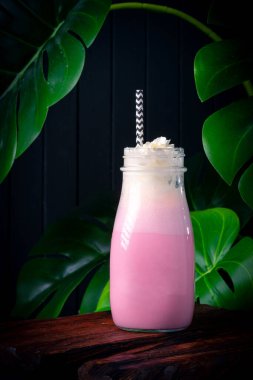 Koyu ahşap arka planda yeşil bitkilerle dolu bir bardakta çilekli milkshake.