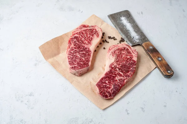 Two raw fat and juicy beef steaks on craft paper with pepper and a meat cleaver