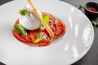 Beyaz tabakta servis edilen fesleğenli ve zeytinyağlı Burrata peyniri ve domates salatası.