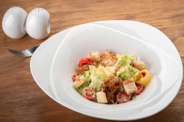 Klasik Sezar Salatası ızgara tavuk, kızarmış ekmek, kiraz domatesleri ahşap bir masada çatalla birlikte beyaz bir tabakta.