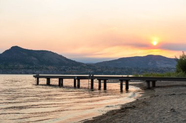 Naramata Merkez Plajı 'ndaki rıhtımın ve sahil şeridinin Naramata, BC, Kanada' daki dağların yaz gün batımı fotoğrafı.