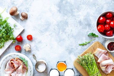Gri beton arka planda yemek pişirmek için gerekli malzemeler. Bir demet taze kuşkonmaz, yumurta, pastırma, mantar, domates, roka. Üst Manzara. Boşluğu kopyala