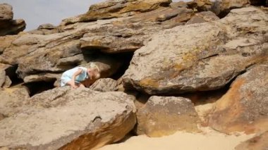 Çocuk taşlara ve taşlara tırmanıyor. Kum taşların arasında görünür. Güzel bir yaz günü. Kız engelleri aşmaktan hoşlanıyor. Sarı saçlı, açık beyaz elbiseli ve sandaletli bir kız.. 