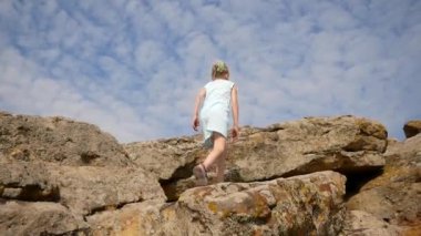 Çocuk taşlara ve taşlara tırmanıyor. Kum taşların arasında görünür. Güzel bir yaz günü. Kız engelleri aşmaktan hoşlanıyor. Sarı saçlı, açık beyaz elbiseli ve sandaletli bir kız.. 