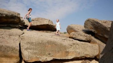 Anne ve çocuk kayalara tırmanıyor, taşlar arasında kum görünüyor. Güzel bir yaz günü. Aile engelleri aşmaktan hoşlanır. Sarı saçlı, açık beyaz elbiseli ve sandaletli bir kız.. 