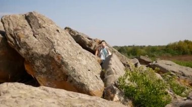 Anne ve çocuk kayalara tırmanıyor, taşlar arasında kum görünüyor. Güzel bir yaz günü. Aile engelleri aşmaktan hoşlanır. Sarı saçlı, açık beyaz elbiseli ve sandaletli bir kız.. 