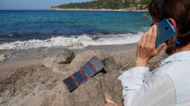 Turist bir kız deniz kıyısında akıllı bir telefonla konuşuyor. Telefon 3 numaralı taşın üzerinde bulunan bir güneş paneli tarafından şarj ediliyor.