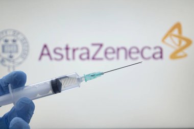 Coronavirus enfeksiyonunu önlemek için Covid-19 aşı şırıngası. Hemşirenin elinde şırınga ve perde arkasında Oxford Üniversitesi Astra-Zeneca logosu