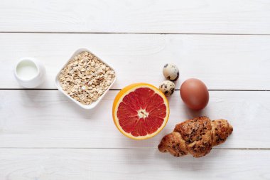 Sağlıklı kahvaltı ürünleri: beyaz ahşap arka planda yulaf ezmesi, greyfurt, yumurta ve kruvasan.