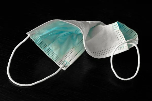 A used medical face mask protecting against respiratory diseases transmitted by airborne droplets such as coronavirus and influenza is crumpled on a black table.