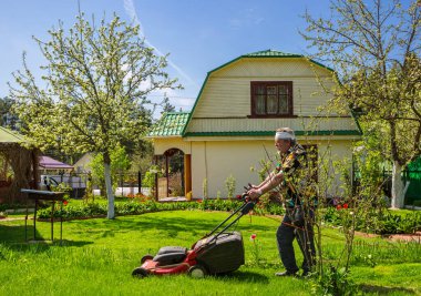 Moscow region, Russia - May 17, 2021:  An elderly man mows the lawn with a lawn mower in the spring at a country house. Light work is good for your health. clipart