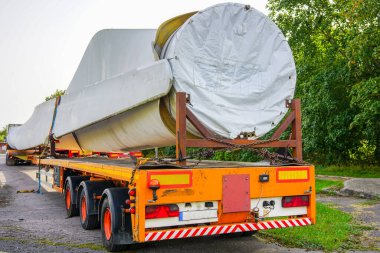 Bir kamyon manzarası, yaz günü otoparkta geniş rüzgar türbini parçaları olan düşük yüklenmiş bir yarı römork.