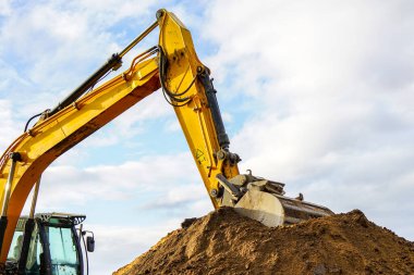 Sarı bir kazıcı, kovayla gökyüzüne karşı toprak yığını kazıyor.