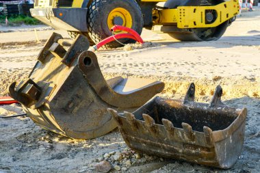 İnşaat alanında iki farklı kazıcı kovası, arka planda sarı traktör.