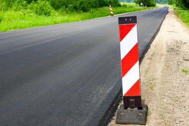 Kırsal kesimde, ön planda kırmızı ve beyaz uyarı işareti olan yeni bir asfalt yol.