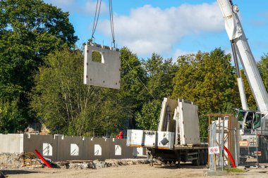 İnşaat alanındaki vinç tarafından kaldırılan ön beton panel. İşçiler, kamyonet ve modern bina montajı sürüyor