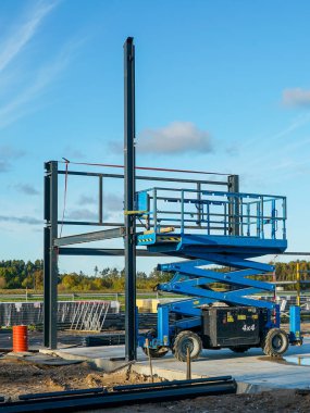 Çelik çerçeve yapısı olan bir inşaat sahasına yerleştirilmiş mavi bir makas kaldıracı. İnşaat, mühendislik ve geliştirme projeleri için endüstriyel ekipman