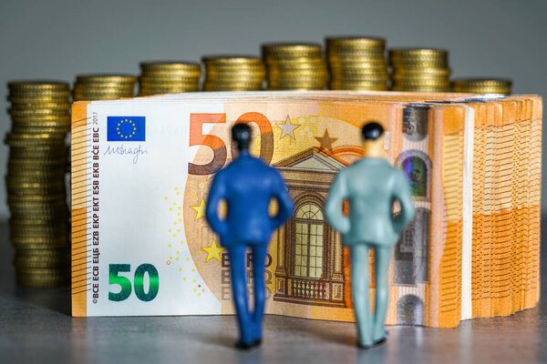 Two miniature businessmen stand in front of tall stacks of golden coins, representing investment, wealth growth and financial success