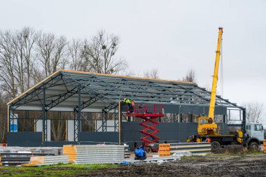 İşçiler, bulutlu bir günde inşaat halindeki çelik çerçeveli sanayi binasının dış sanayi duvarlarını örmek için makas asansörü ve vinç kullanıyorlar.