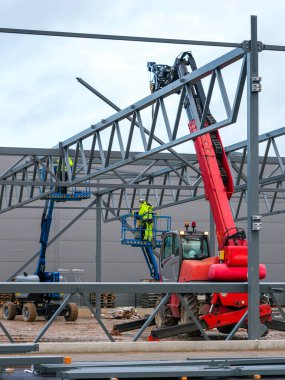 Güvenlik donanımlı inşaat işçileri, endüstriyel bir inşaat alanında vinç ve hava asansörleri kullanarak çelik bir iskelet oluşturuyorlar.