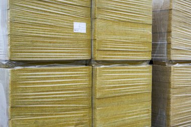 Plastiğe sarılı ve dışarıda istiflenmiş, endüstriyel bir inşaat alanında kullanıma hazır mineral yün yalıtım tahtaları.