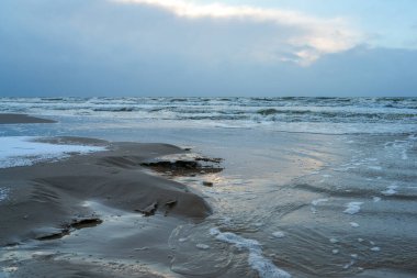 Baltık Denizi kıyısında soğuk bir kış akşamı dalgalar, ıslak yansıtıcı kum, deniz köpüğü ve alacakaranlıkta karamsar bir gökyüzü.