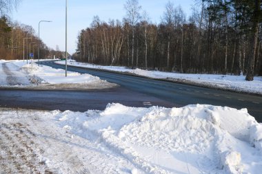Orman ağaçları, asfalt ve mavi gökyüzünün altındaki kar yığınları ile karlı bir kış manzarasında kıvrılan sakin bir banliyö yolu.