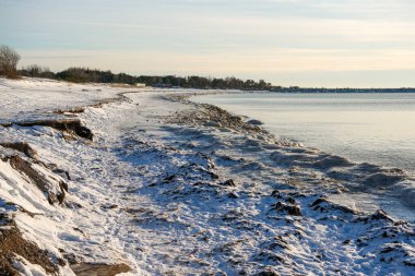 Letonya 'nın Liepaja kentindeki Baltık Denizi kıyılarında kış mevsimi, kar kaplı kum tepeleri, buzlu kıyı şeridi ve yumuşak ışık altında sakin su