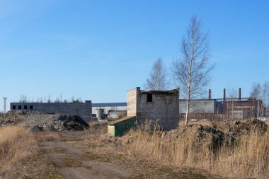 Eski tuğla bina, kuru ot, toprak yol ve temiz gökyüzü altındaki uzak fabrika yapılarıyla ıssız bir sanayi bölgesi.