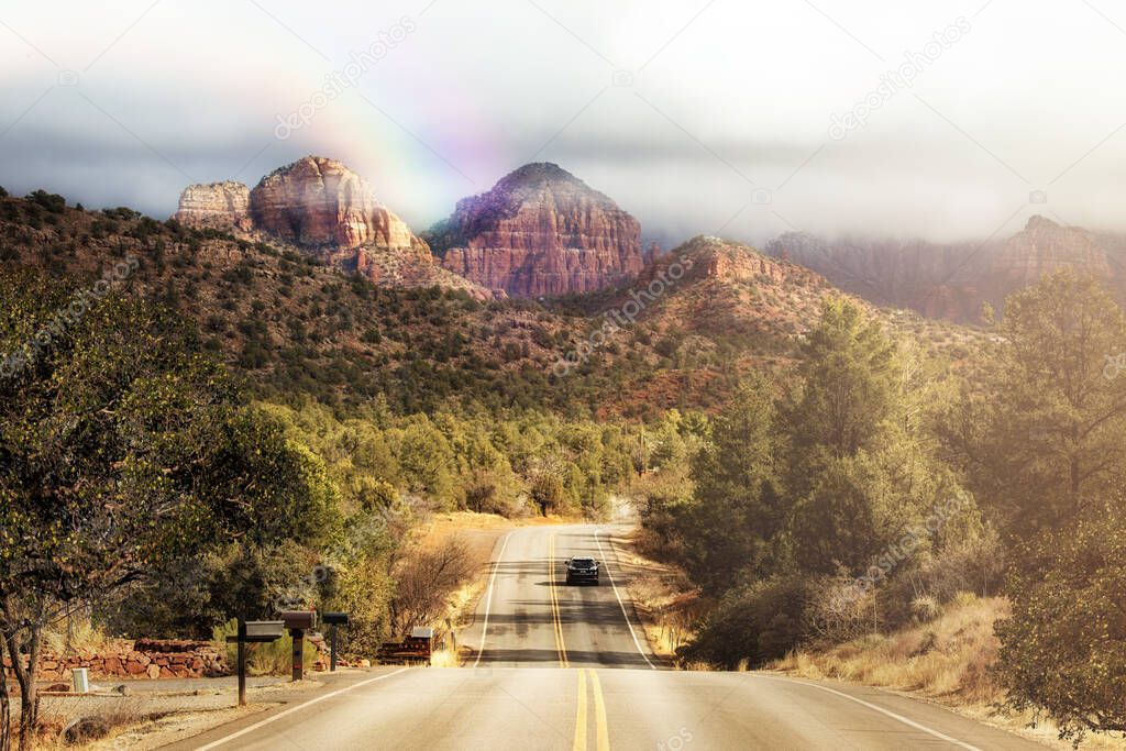 Road trip down a winding Chavez Ranch Road leading to Chapel Rock and Catherdal Rock in the northern Arizona town of Sedona with dramatic clouds and a rainbow in the sky.