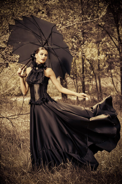 Magnificent brunette woman wearing long black dress walking in a mystic forest. The old times, the Gothic style. Fashion.