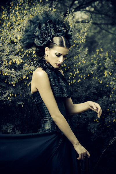 Gothic brunette woman in long black dress and beautiful black headwear posing in the blossoming garden. Medieval, old times. Fashion. Gothic style.