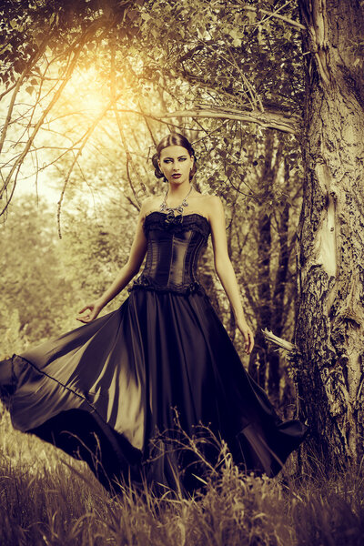 Magnificent brunette woman in black old-fashioned dress walking in the thicket of the magic forest. Gothic style. Fashion.