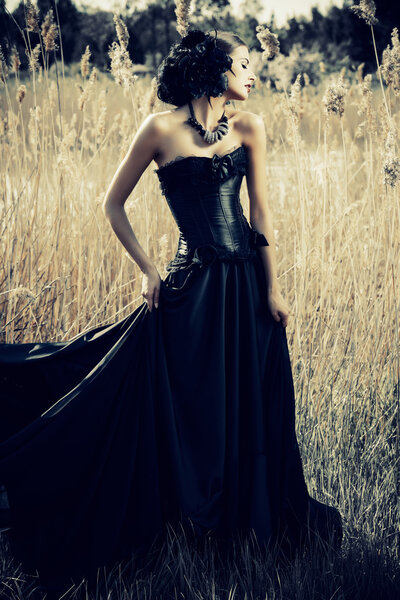 Beautiful brunette woman wearing long black dress and black headwear posing among the reeds. The old times, the Gothic style. Fashion.