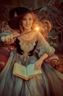 Şapkalı ve güzel elbiseli büyüleyici bir cadının portresi bir büyü kitabı ve eski kalede sihirli bir değnekle koltukta oturuyor. Cadılar Bayramı. Kutlama.