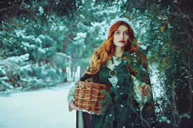 Portrait of a beautiful young woman with long red hair wearing medieval clothes walks through a winter snowy forest. Historical reconstruction of the early Middle Ages. Christmas tale. clipart
