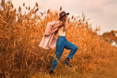 Sonbahar modası. Güz manzarasının arka planında sazlıkların yanında poz veren modaya uygun giyinmiş güzel bir genç kadının tam boy portresi..