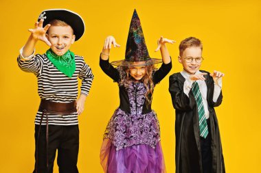 Cadılar Bayramı 'nda şeker mi şaka mı? Cadılar Bayramı kostümlü üç neşeli çocuk stüdyoda sarı bir arka planda duygusal poz veriyor. Boşluğu kopyala. 