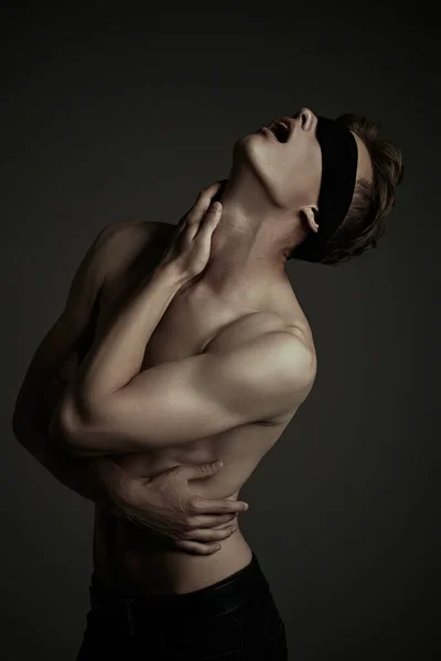 Art portrait of a handsome blindfold young man with perfect muscular body raised his head up, expressing tension and suffering. Black background, darkness.
