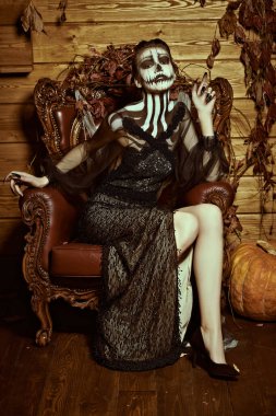 Balkabağı makyajlı zarif bir bayan sonbahar süslemelerinde klasik koltukta oturur. Cadılar Bayramı. Ölülerin Günü. 