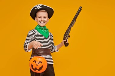 Mutlu Cadılar Bayramı! Stüdyoda sarı arka planda balkabağı sepeti ve silahla poz veren korsan kostümlü neşeli çocuk. Boşluğu kopyala. 