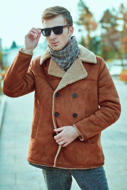 Erkek tarzı. Kışlık ceketli ve atkılı yakışıklı genç adam kare şeklinde duruyor. Moda, giysi ve aksesuarlar..